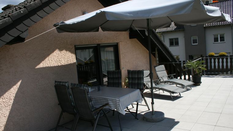 Eine sonnige Terrasse mit einem Tisch, Stühlen und Sonnenliegen. Ein Sonnenschirm sorgt für Schatten.