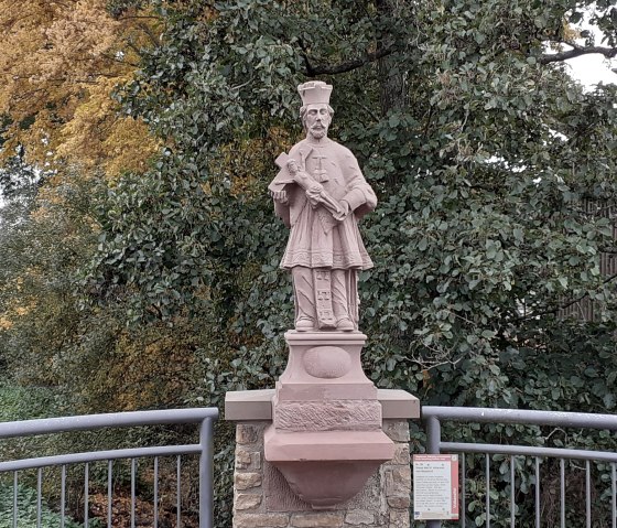 Statue des hl. Johannes von Nepomuk auf einer Br&uuml;cke, umgeben von herbstlichen B&auml;umen und einem Gel&auml;nder., &copy; Touristik GmbH Gerolsteiner Land