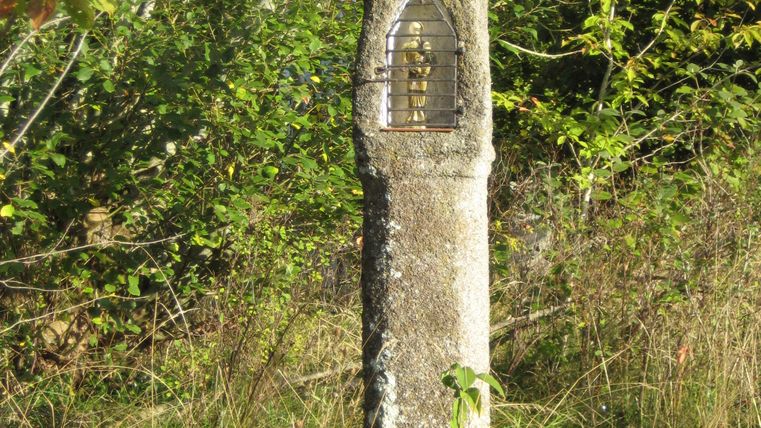 Steinernes Wegekreuz mit kleinem Dach und Gitter, umgeben von Bäumen und Gras.