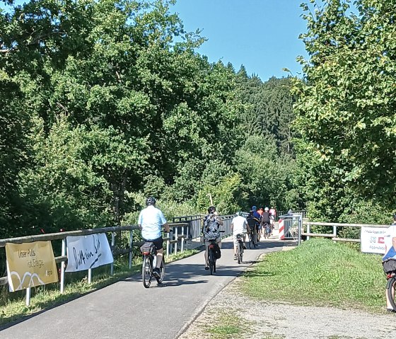 Mehrere Personen auf Fahrrädern radeln über den geteerten Kyll-Radweg bei Stadtkyll. Am Zaun neben dem Radweg hängen Banner von Sponsoren.