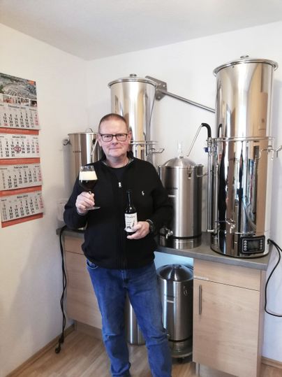 Ein Mann steht vor einer Brauanlage und hält eine Flasche Bier und ein gefülltes Glas hoch. 