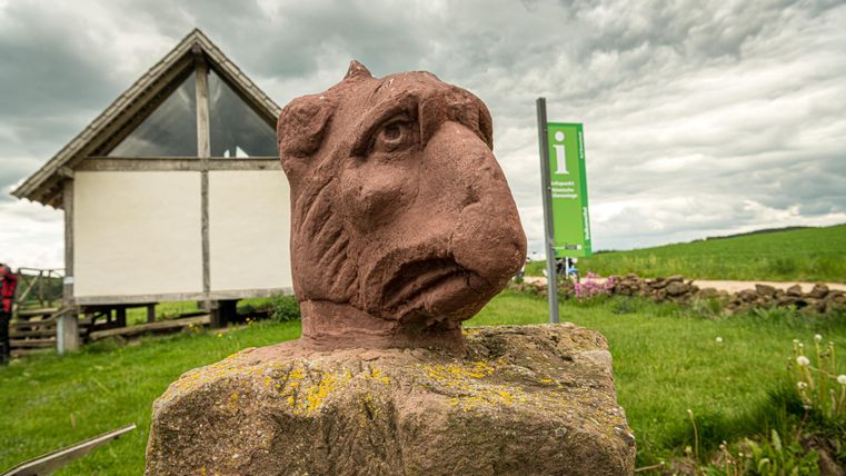 Steinskulptur eines Greifenkopfes vor einem kleinen Gebäude und einer Infotafel auf einer Wiese unter bewölktem Himmel.