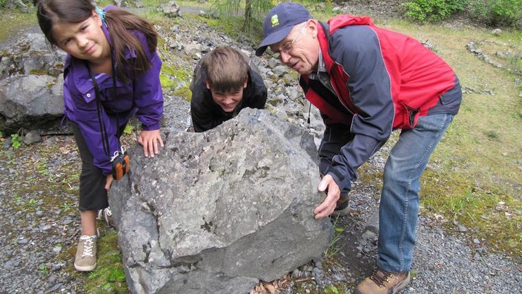 Ein Mädchen und ein Junge stehen neben einem großen Stein und versuchen, ihn zu heben. Ein älterer Mann hilft ihnen dabei und lächelt.