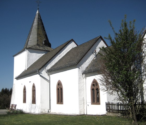 Witte kerk met een spitse toren en gotische ramen, omringd door bomen en weiden, onder een helderblauwe hemel., © Touristik GmbH Gerolsteiner Land