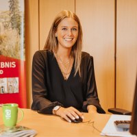Eine Frau sitzt an einem Schreibtisch und l&auml;chelt in die Kamera. Hinter ihr einige Schr&auml;nke und ein Banner mit Aufdruck "Freizeirerlebnis Gerolsteiner Land" und Bild einer Burg.