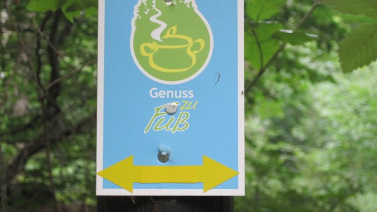 Schild mit Logo 'Genuss zu Fuß' und gelbem Pfeil nach links im Wald.
