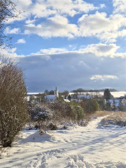 Eine verschneite Landschaft mit einem kleinen Dorf und einer Kirche im Hintergrund. Der Himmel ist teilweise bewölkt und verbreitet eine ruhige Winteratmosphäre.