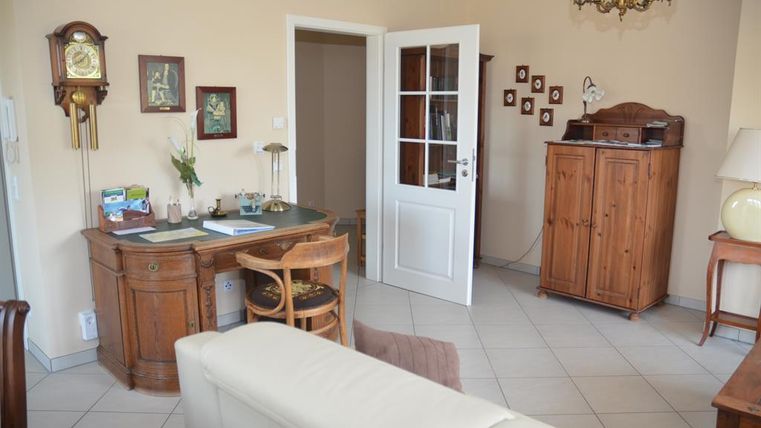 Een gezellige woonkamer met een bureau, een stoel en een houten commode. De muren zijn in een warme kleur en er zijn enkele schilderijen en planten.