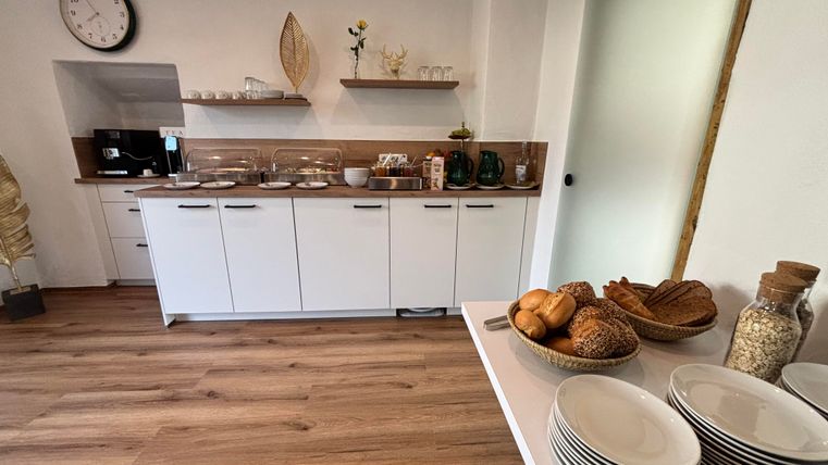 Eine moderne Küche mit weißen Möbeln und Holzregalen. Auf dem Tisch sind Geschirr und frische Backwaren angerichtet.