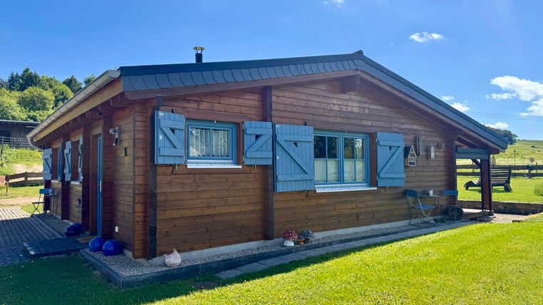 Ein gemütliches Holzhaus mit blauen Fensterläden, umgeben von grünem Gras. Der Himmel ist klar und sonnig.