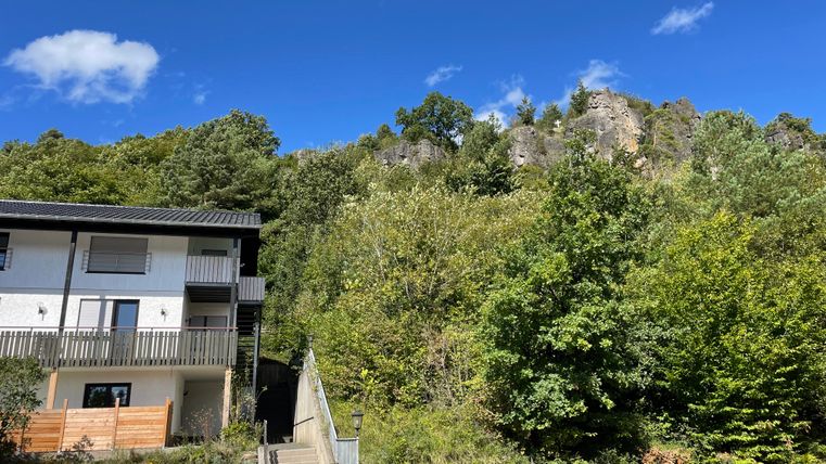 Un bâtiment moderne à côté d'une verdure luxuriante et d'arbres. À l'arrière-plan, on voit des falaises escarpées et un ciel bleu clair.