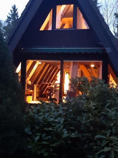 Ein gemütliches Ferienhaus mit einem spitzen Dach, beleuchtet von innen. Umgeben von grünen Pflanzen in der Dämmerung.