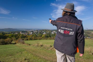 Une personne portant un chapeau et une veste avec l'inscription 'auf EIFEL KRIMI SPUR' montre un paysage avec un village en arrière-plan.