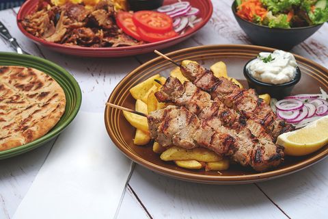 Sur une table en bois, il y a quatre plats : gyros avec frites, viande, pain et une salade mixte.