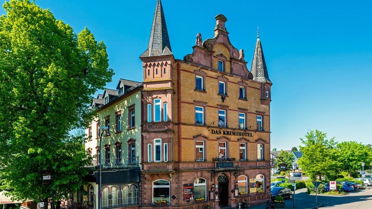 Historisches Gebäude mit Türmen und der Aufschrift 'Das Krimihotel', umgeben von Bäumen und blauen Himmel.