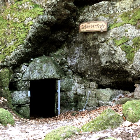 Ein moosbedeckter Felsen mit einem dunklen Höhleneingang, beschriftet mit 'Schwedenfeste'. Herbstlaub bedeckt den Boden., © Touristik GmbH Gerolsteiner Land