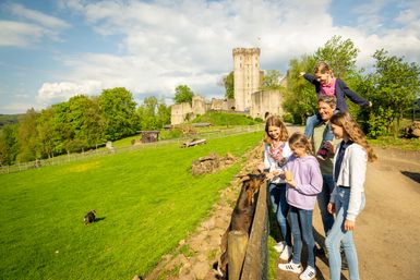 Eine Familie füttert eine Ziege vor einer Burg. Im Hintergrund grüne Wiesen und Bäume, der Himmel ist leicht bewölkt.