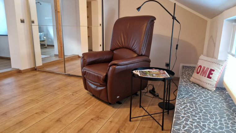 Ein gemütlicher Sessel aus braunem Leder steht in einem hellen Raum mit Holzboden. Neben dem Sessel befindet sich ein kleiner Tisch mit einer Leselampe und einem Kissen mit der Aufschrift „HOME“.