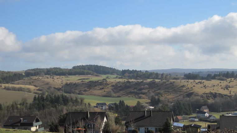 Landschaft mit Hügeln, Wiesen und Häusern unter blauem Himmel.