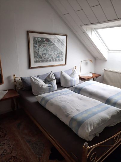 Ein gemütliches Schlafzimmer mit zwei Betten und weißen Bettdecken. Ein Fenster lässt Tageslicht herein und ein Gemälde hängt an der Wand.