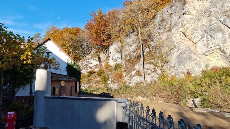 A picturesque house at the foot of a rock wall, surrounded by colorful autumn trees. The clear sky and the natural surroundings create a tranquil atmosphere.