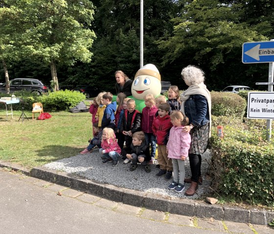 Statue von Maskottchen Willi Basalt mit grünem Shirt umgeben von einigen Kindergarttenkindern und ihren Erzieherinnen.