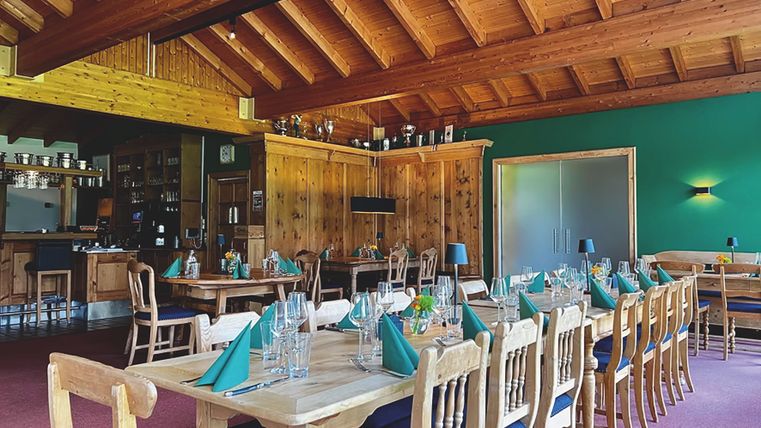In einem Raum mit Holzdecke steht eine lange Holztafel, die in der Farbe grün eigededeckt ist, Im Hintergrund sind weitere gedeckte Tische