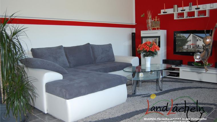 Ein modernes Wohnzimmer mit einer grauen Couch und einem Glastisch. Die Wände sind in Rot und Weiß gestrichen, mit dekorativen Elementen und einem Fernseher.