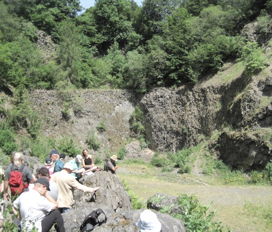 Eine Gruppe von Menschen sitzt auf Felsen in einer gr&uuml;nen, felsigen Landschaft. Im Hintergrund sind B&auml;ume und steile Felsw&auml;nde zu sehen., &copy; Touristik GmbH Gerolsteiner Land, Ute Klinkhammer