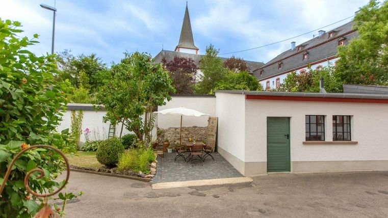 Ein ruhiger Innenhof mit einem kleinen Tisch und Stühlen, umgeben von Bäumen und Blumen. Im Hintergrund ist ein Kirchturm sichtbar.