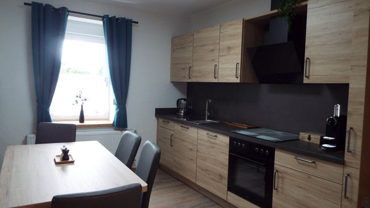 Eine moderne Küche mit Holzschränken und einem Esstisch. Vor dem Fenster hängen dunkle Vorhänge, und die Küchenzeile ist gut ausgestattet.