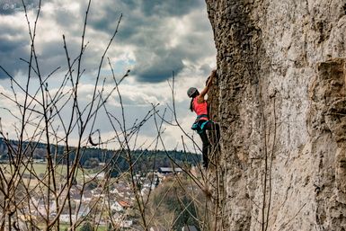 Eine Person klettert an einer steilen Felswand. Im Hintergrund ist ein Dorf und bewölkter Himmel zu sehen.