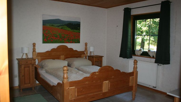 Een gezellige slaapkamer met een houten tweepersoonsbed. De kamer heeft een raam met groene gordijnen en een kleurrijk schilderij aan de muur.