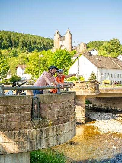 Von der Brücke aus blickt man auf einen Fluss; im Hintergrund stehen eine Burg auf einem Hügel und Häuser, im Vordergrund sitzt eine Person auf der Brückenmauer.