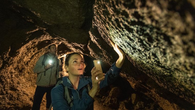 Zwei Personen in einer Höhle, beleuchten die Wände mit Taschenlampen. Eine Person hält ein Smartphone, die andere eine Taschenlampe.