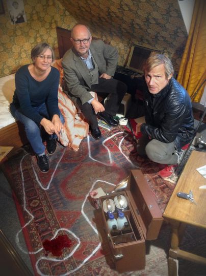 Drei Personen in einer Dachkammer neben einem Kreideumriss und einer Blutlache auf einem Teppich. Ein offener Koffer mit Flaschen steht daneben.