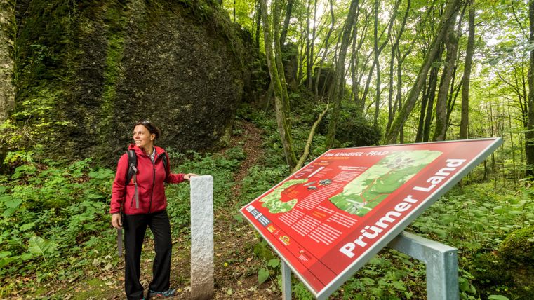Eine Frau in roter Jacke steht neben einem Informationsschild im Wald.