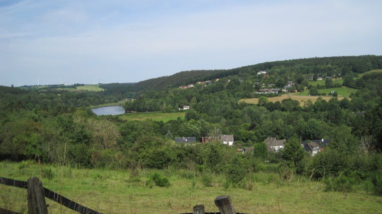 Landschaft mit Blick auf den Kronenburger See, umgeben von grünen Hügeln und Wäldern, im Vordergrund ein Holzzaun.