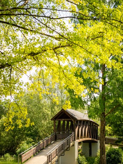 Eine überdachte Holzbrücke führt über einen Fluss in einem stark blühenden Park.