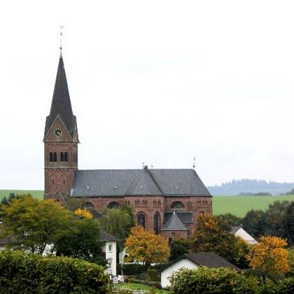Eine Kirche mit hohem Turm in einer ländlichen Umgebung, umgeben von Bäumen und Häusern.