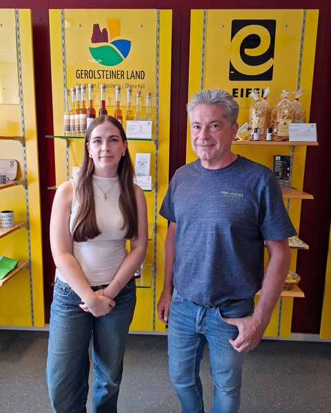Ein Mann und eine junge Frau stehen nebeneinander vor eine Shop-Wand und lächeln in die Kamera.