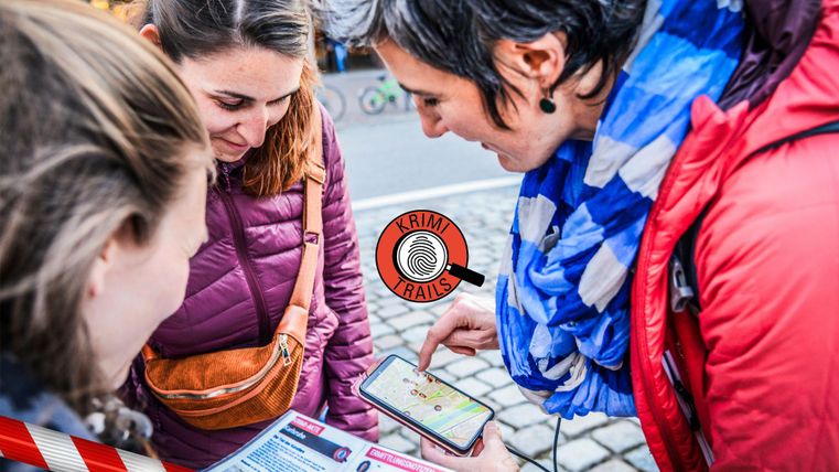 Drei Frauen schauen auf ein Smartphone und diskutieren gemeinsam. Im Hintergrund sind Stadtansichten zu erkennen, die das urbane Umfeld zeigen.