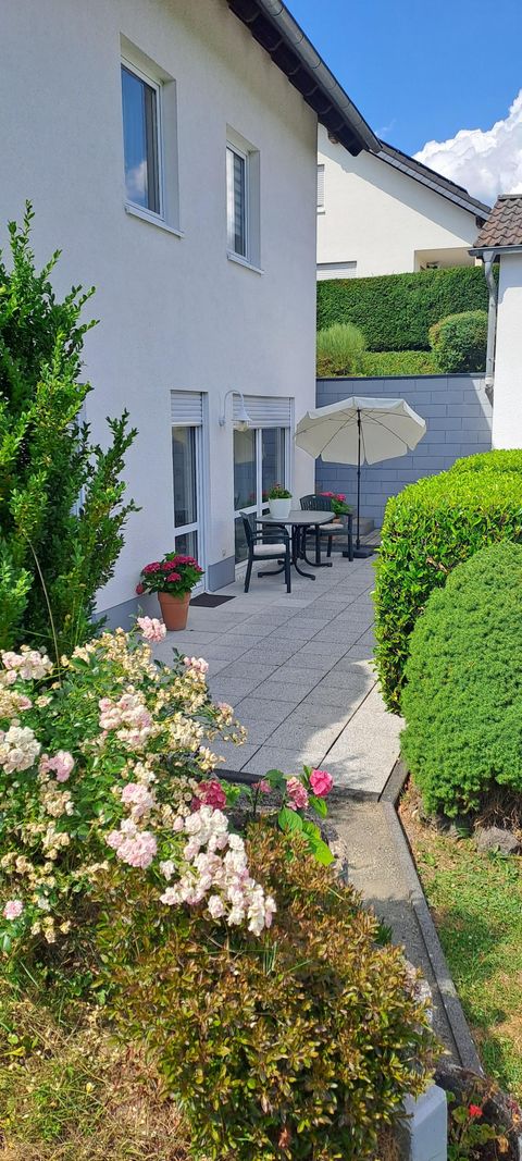 A cozy terrace with flowers and a sun umbrella. In the background, a white house and well-kept hedges are visible.