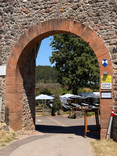 Ein Durchgang durch einen steinernen Torbogen führt zu einem bewirteten Außenbereich mit Tischen und Schirmen. An der rechten Säule des Torbogens befinden sich Schilder, die darauf hinweisen, dass die Durchfahrt verboten ist und eine geschlossene Veranstaltung stattfindet.