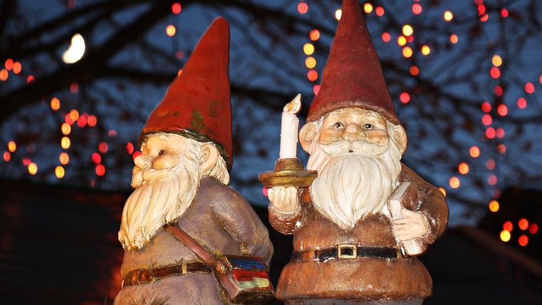 Two decorative garden gnomes, one of whom is holding a candle. Colorful lights are glowing in the background.