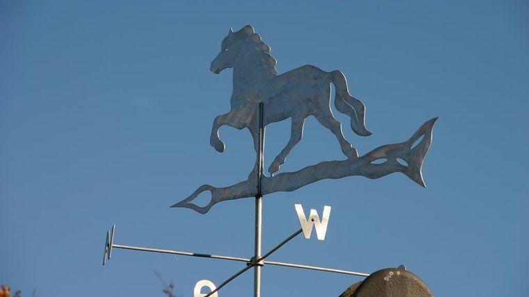Een windspel in de vorm van een paard op een dak. De lucht is helder en blauw.