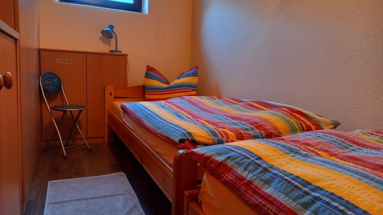 A cozy bedroom with two beds and colorful bedding. There is a small desk, a chair, and a window that lets in daylight.