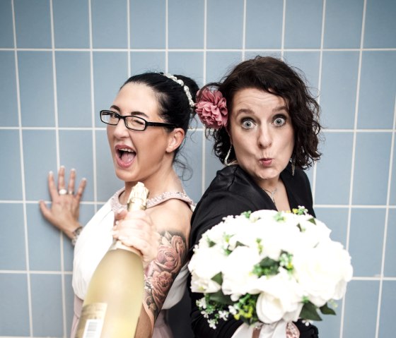 Two women in stylish clothing pose side by side with a bottle of sparkling wine and a bouquet of flowers in their hands.