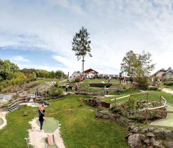 Panoramabild eines Minigolfplatzes in grüner Landschaft. Mehrere Personen spielen auf verschiedenen Bahnen. Im Hintergrund sind Bäume und ein Gebäude zu sehen., © Eifel-Adventure, Andreas Goltz