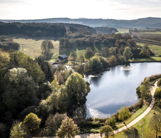 Luftaufnahme eines Sees im Bolsdorfer Tälchen, umgeben von Wäldern und Feldern. Ein Weg führt entlang des Ufers. Die Landschaft ist herbstlich gefärbt., © Eifel Tourismus GmbH, D. Ketz